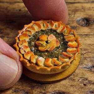 Può includere: Una torta salata rotonda in miniatura con un bordo di crosta dorata e ondulata e un centro verde scuro alle erbe, guarnita con una disposizione circolare di sottili fette di patate dorate, presentata come una replica di cibo.