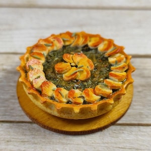 May include: A miniature round savory tart featuring a golden-brown scalloped crust. The filling consists of dark green cooked greens topped with a circular arrangement of thinly sliced, roasted golden-brown root vegetables and green herbs.