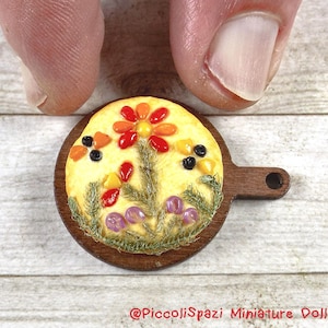 Op de afbeelding: Miniatuur houten snijplank met een bloemenontwerp. Het gele oppervlak heeft een bloem met rode en oranje bloemblaadjes en andere kleurrijke bloemen en bladeren. De plank heeft een handvat en is ongeveer 2,5 cm in diameter.