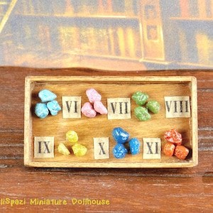 Può includere: Un vassoio di legno con numeri romani da IX a VIII, ogni numero ha un piccolo mucchio di pietre colorate. Le pietre sono blu, verdi, gialle, rosa e rosse.