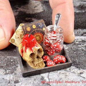 May include: Miniature Halloween display with a skull and red candle, a jar of red and white spotted mushrooms, and small books on a wooden tray. The scene is set against a textured grey surface.