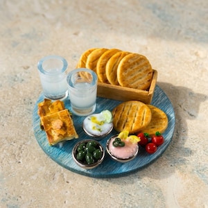 Può includere: Una presentazione di cibo in miniatura su un vassoio circolare blu con due bicchieri trasparenti, una cassetta di legno con cracker rotondi, pezzi di pasta quadrati, una ciotola di olive verdi, una ciotola di salsa bianca e una salsa rosa.