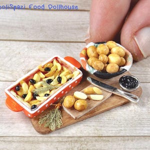 Può includere: Scena di cibo in miniatura con un piatto di spicchi di patate e olive nere, una piccola ciotola di patate e un tagliere di legno con un coltello. Su una superficie di legno chiaro.
