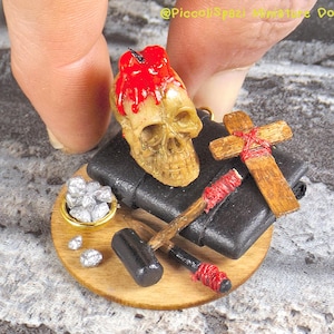 May include: A miniature dollhouse scene featuring a skull candle, a small wooden cross, a hammer, a small bowl of silver coins, and a black book on a wooden base.