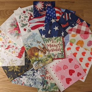 May include: A collection of colorful paper napkins with various designs, including floral patterns, patriotic themes, and holiday greetings. The napkins feature images of butterflies, rabbits, hearts, and stars. Some napkins have text, such as "Merry Christmas" and "Happy Holidays".