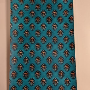 May include: A teal blue fabric with a repeating pattern of a muscular man in a black and orange circle. The man is holding a weight in each hand and has a determined expression on his face. The text "Iron Maiden" is written in black letters below the circle.