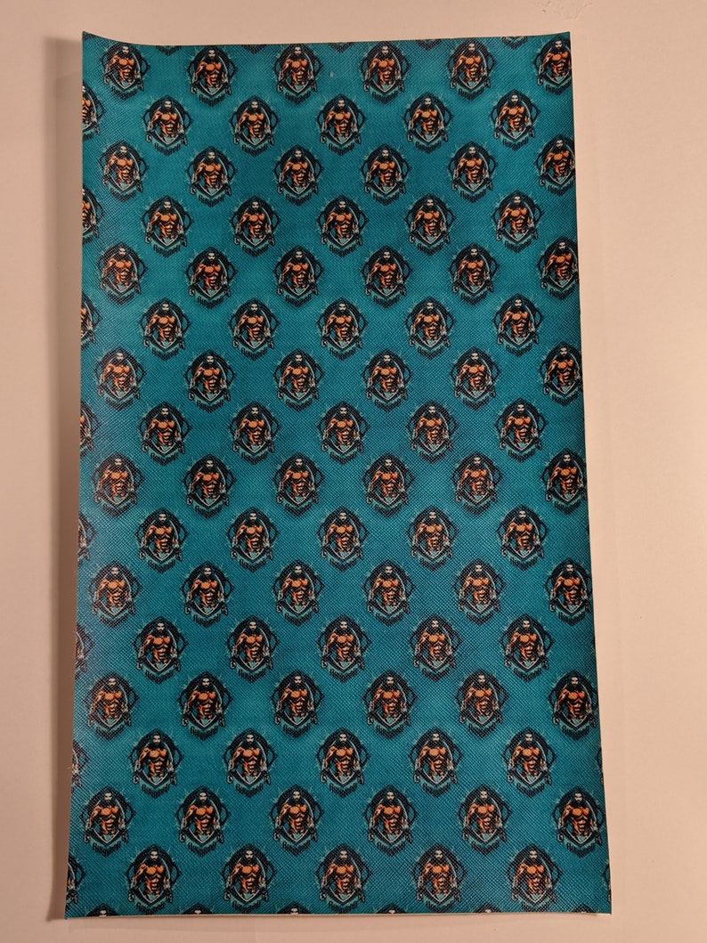 May include: A teal blue fabric with a repeating pattern of a muscular man in a black and orange circle. The man is holding a weight in each hand and has a determined expression on his face. The text "Iron Maiden" is written in black letters below the circle.