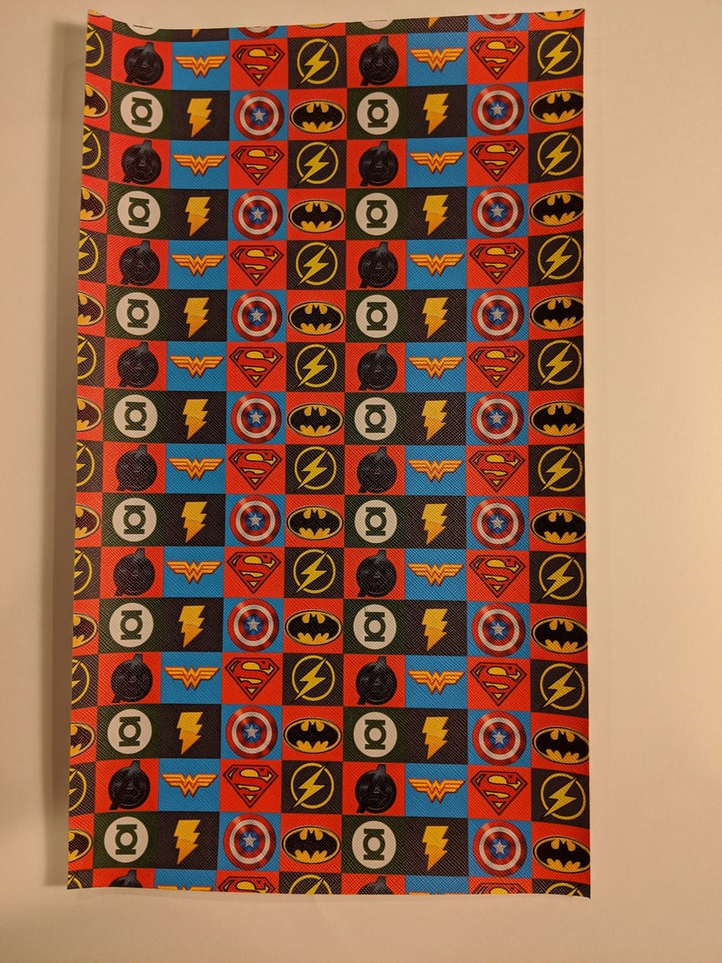 May include: A sheet of wrapping paper with a repeating pattern of superhero logos. The logos include Superman, Batman, Wonder Woman, The Flash, and Green Lantern. The logos are in a checkered pattern of red, blue, yellow, and black.