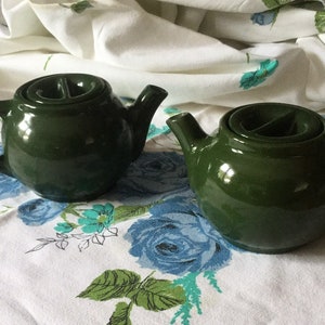 May include: Two green ceramic teapots with lids. The teapots have a unique design with a small opening in the lid.