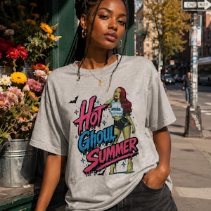 May include: A heather gray short-sleeve t-shirt featuring a graphic print of a stylized character with red hair and green skin, wearing a white crop top and black shorts. The text "Hot Ghoul SUMMER" is printed in pink and blue with a dripping effect.