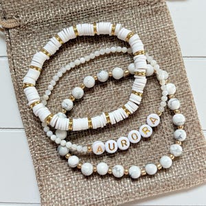 May include: Four white and gold beaded bracelets, one spelling "AURORA". The bracelets are presented on a small, tan burlap drawstring bag. The beads are in various sizes.