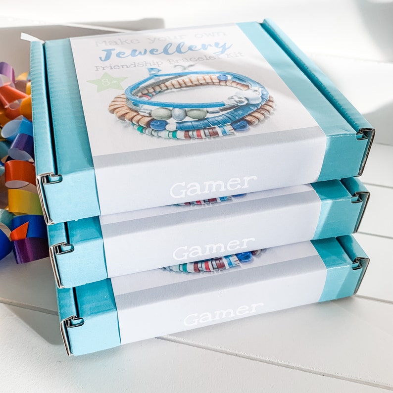 Stack of three blue boxes with white labels. The top box has a label that reads 'Make Your Own Jewellery Friendship Bracelet Kit 8+' and shows a colorful friendship bracelet. The other two boxes have labels that read 'Gamer'.