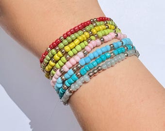May include: A collection of beaded bracelets in various colors, including red, green, yellow, pink, blue, turquoise, and gray. Each bracelet features small, colorful beads and silver-colored accent beads. The bracelets are stacked on a wrist.