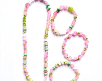May include: A long necklace and three circular bracelets made of alternating round beads in shades of translucent pink, opaque light green, and clear white. The jewelry set features a repeating pattern of these colorful beads strung together.