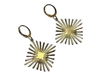 May include: A pair of gold-colored earrings. Each earring features a square design with a sunburst pattern radiating from a central circle. The earrings have a lever-back closure and are set against a white background.