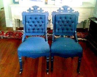 May include: Two matching blue chairs with tufted backrests and ornate wooden frames. Each chair features a high back with decorative carved detailing at the top, upholstered seats, and small wheels attached to the bottom of the four wooden legs.