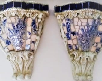 May include: A pair of wall-mounted sconces featuring a cream-colored base with ornate carved details. The front surfaces are decorated with a mosaic pattern of blue and white tiles, including floral motifs and a row of dark blue square tiles.
