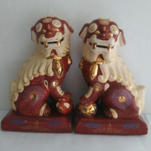 May include: A pair of ceramic Foo dog statues featuring a deep red body with cream-colored manes and tails. Each figure is accented with gold detailing on the face, paws, and base, and includes a small spherical object positioned under one front paw.