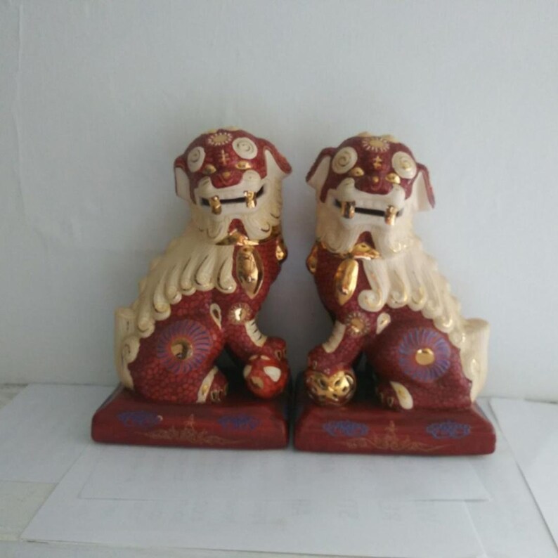 May include: A pair of ceramic Foo dog statues featuring a deep red body with cream-colored manes and tails. Each figure is accented with gold detailing on the face, paws, and base, and includes a small spherical object positioned under one front paw.