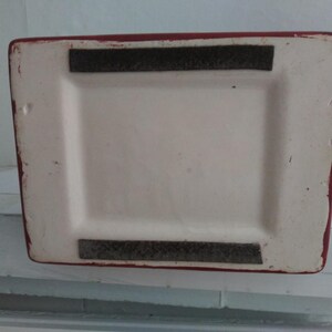 May include: The rectangular base of a ceramic object features a white center with a recessed area. Two parallel, dark gray felt strips are attached to the top and bottom edges of the white surface. The outer border of the base is painted red.