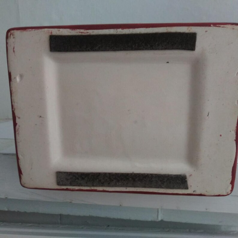 May include: The rectangular base of a ceramic object features a white center with a recessed area. Two parallel, dark gray felt strips are attached to the top and bottom edges of the white surface. The outer border of the base is painted red.