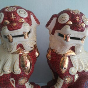 May include: A pair of ceramic Foo Dog statues featuring a deep red textured body with cream-colored manes and tails. The figures are accented with metallic gold details on the eyes, teeth, paws, and swirling patterns across the heads and chests.