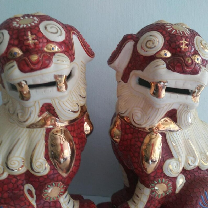 May include: A pair of ceramic Foo Dog statues featuring a deep red textured body with cream-colored manes and tails. The figures are accented with metallic gold details on the eyes, teeth, paws, and swirling patterns across the heads and chests.