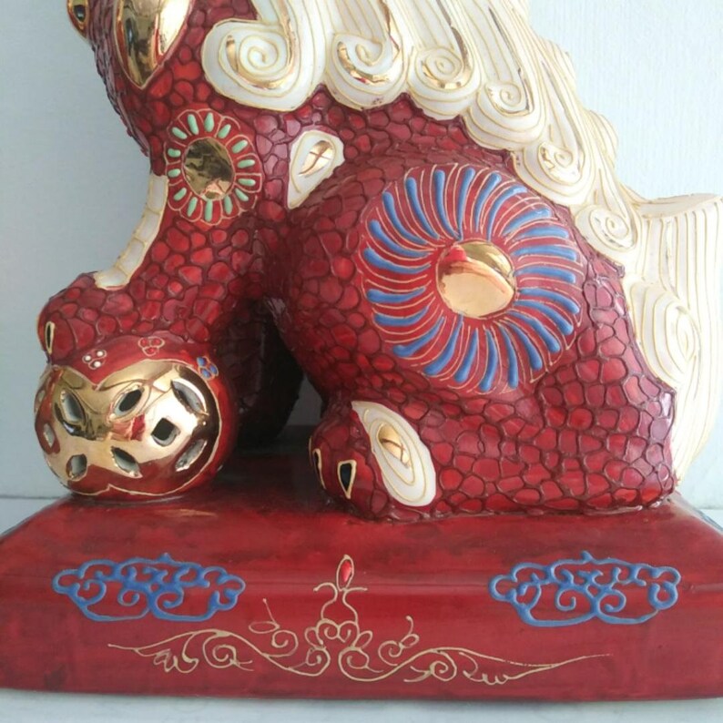 May include: A ceramic guardian lion statue featuring a textured red body with gold, white, and blue accents. The figure rests on a rectangular red base decorated with blue cloud motifs and gold filigree, with one paw resting on a gold sphere.