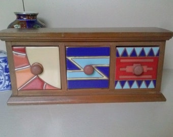 May include: A wooden desktop storage unit with three drawers featuring ceramic fronts. The left drawer has a cream and red geometric design, the center drawer has a blue and light blue zigzag pattern, and the right drawer has a red and blue triangle motif.