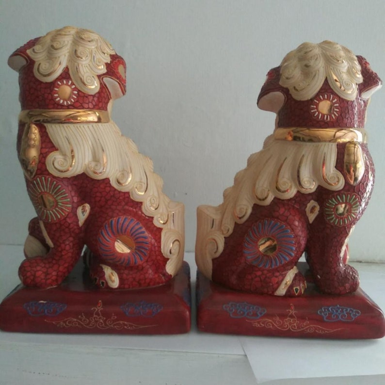 May include: A pair of ceramic Foo Dog statues featuring a textured red body with cream-colored manes and tails. Each figure has gold-painted collars and accents, with blue and gold circular patterns on the legs, standing on rectangular red bases.
