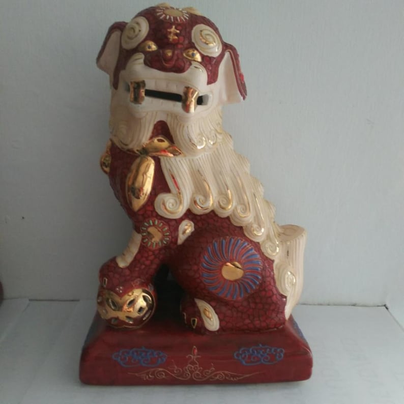 May include: A ceramic Foo Dog figurine featuring a deep red body with a crackle glaze finish, accented by cream-colored mane and tail details. The creature has gold-painted highlights, a wide mouth with a coin slot, and rests on a rectangular base.