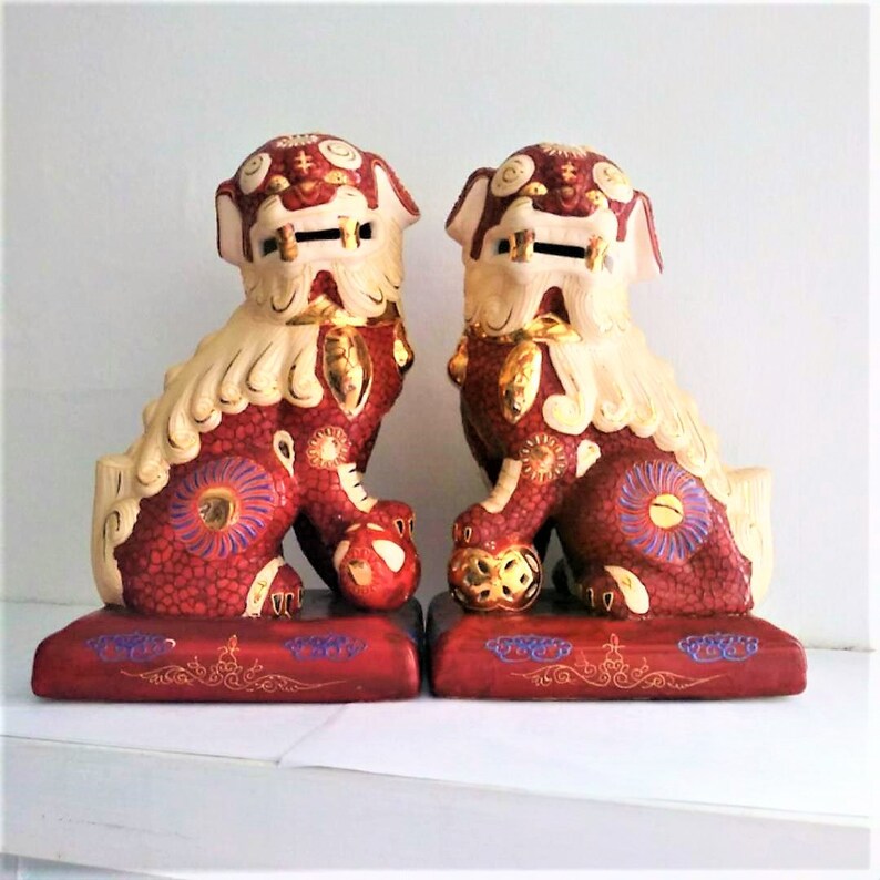 May include: A pair of ceramic Foo Dog statues featuring a deep red textured body with cream-colored manes and gold accents. Each guardian lion sits on a rectangular red base, with one paw resting on a decorative ball, showcasing intricate painted details.