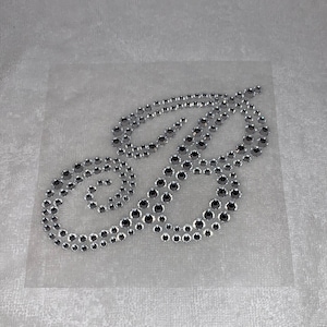 May include: A silver rhinestone design in the shape of the letter "B" on a clear adhesive sheet.