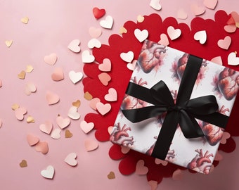 May include: A square gift box wrapped in white paper featuring a repeating anatomical heart pattern in shades of red and pink. The box is tied with a wide, smooth black satin ribbon tied into a bow on the top center of the package.