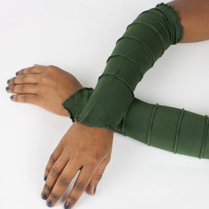 May include: Dark green arm warmers with a ruffled edge and stitched seams. The arm warmers are shown on a white background. The sleeves are long and cover the arms from the wrist to the upper arm.