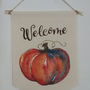 May include: A canvas banner with a painted pumpkin and the word "Welcome" in black script. The pumpkin is orange, red, and blue. The banner is suspended by a wooden dowel and twine.