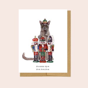May include: A gray cat wearing a crown sits in front of three nutcracker figurines. The cat is looking at the viewer. The nutcrackers are wearing red and gold uniforms. The text "Prrrrrr Rum Pum Pum Pum" is written below the image.