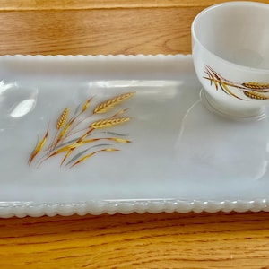 Puede incluir: Un juego de merienda de vidrio lechoso blanco que consta de una bandeja rectangular con bordes festoneados y asas, junto con una taza a juego. Ambas piezas presentan un patrón decorativo de tallos de trigo dorados y marrones.