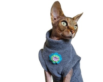 May include: A gray cat wearing a gray sweater with a blue and green flower on the front. The cat is sitting on a white background.