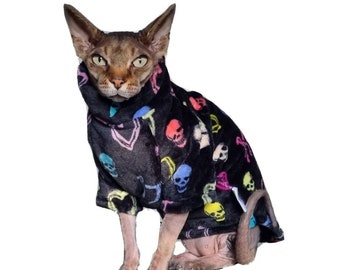 Puede incluir: Un gato negro con un suéter de vellón negro con un patrón de calaveras de colores. El suéter tiene cuello alto y mangas largas.