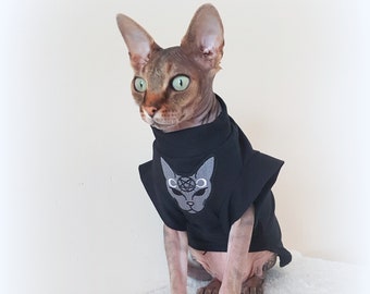 May include: A hairless cat wearing a black t-shirt with a gray cat face and pentagram design. The cat is sitting on a white fluffy surface.