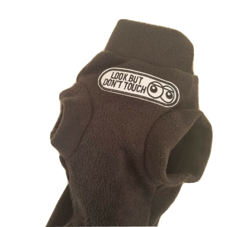 May include: A dark brown fleece garment with a high collar and arm openings. A white patch with the text "LOOK BUT DON'T TOUCH" and cartoon eyes is sewn on the collar. The garment is likely for a pet.