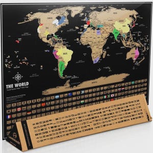 May include: A black scratch-off world map with gold-colored landmasses, featuring colorful country highlights, a compass rose, and the text THE WORLD Travel Tracker Map with Flags. A triangular cardboard tube with flag icons sits in the foreground.