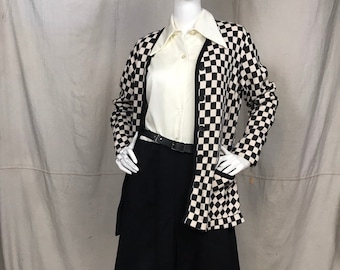 May include: A mannequin displays a vintage outfit. The ensemble includes a cream-colored button-down shirt, a black and cream checkered cardigan, a black belt, and black knee-length shorts. The cardigan has pockets and a black trim.