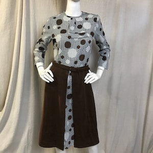 Op de afbeelding: Een vintage jurk met een grijs en wit polkadotpatroon op de top en mouwen, met grote bruine polkadots. De rok is effen bruin van kleur en de jurk heeft een bruine riem. De jurk wordt op een mannequin gepresenteerd.