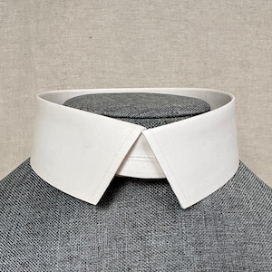 以下が含まれることがあります： 尖った先端と幅広の平らなバンドが付いた白いドレスシャツの襟。襟は灰色のマネキンに着せられています。