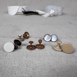 May include: A collection of various shirt studs and buttons, including a white circular button, a pair of iridescent mother-of-pearl studs, a beige button, and several metallic bronze-toned studs with stems, arranged on a textured fabric surface.