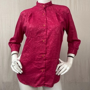 May include: A long-sleeved button-down shirt in a vibrant magenta color featuring a tonal floral jacquard pattern throughout. The shirt has a mandarin collar, a full front button placket with matching buttons, and cuffed sleeves.