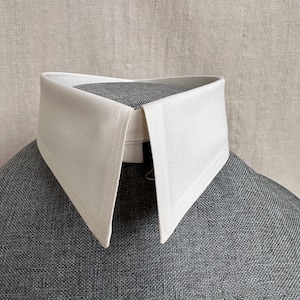 May include: A crisp, off-white detachable shirt collar featuring a structured, pointed design with visible fabric texture. The collar is displayed on a grey, textured form, showcasing its clean lines, stiff construction, and classic shape.