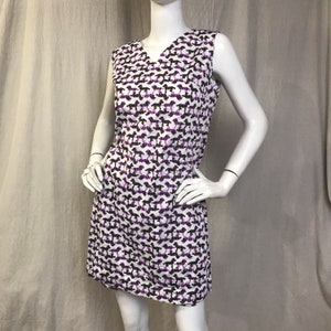 May include: Sleeveless white dress with a V-neck, patterned with black and purple dog silhouettes. The dress is knee-length and has a simple, classic design. The dress is displayed on a mannequin.